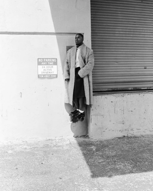 A black and white portrait of an adult male wearing a long coat and roller skates, leaning against a wall with signage.