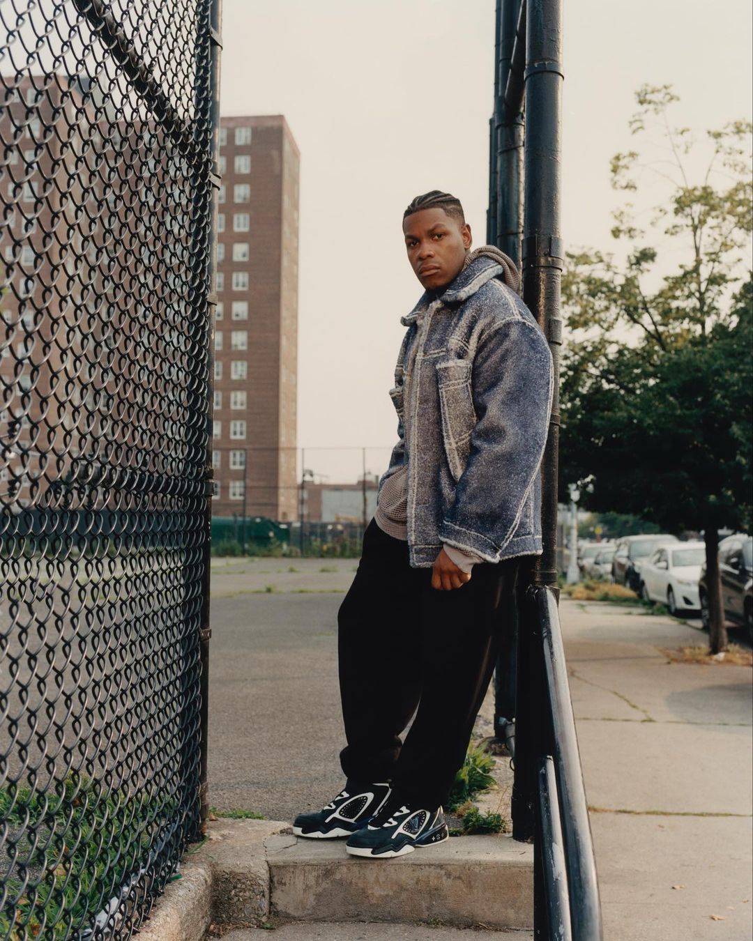 Eye-level outdoor shot of an adult male standing on concrete steps, wearing a jacket, hoodie, black pants and sneakers, with a metal fence.