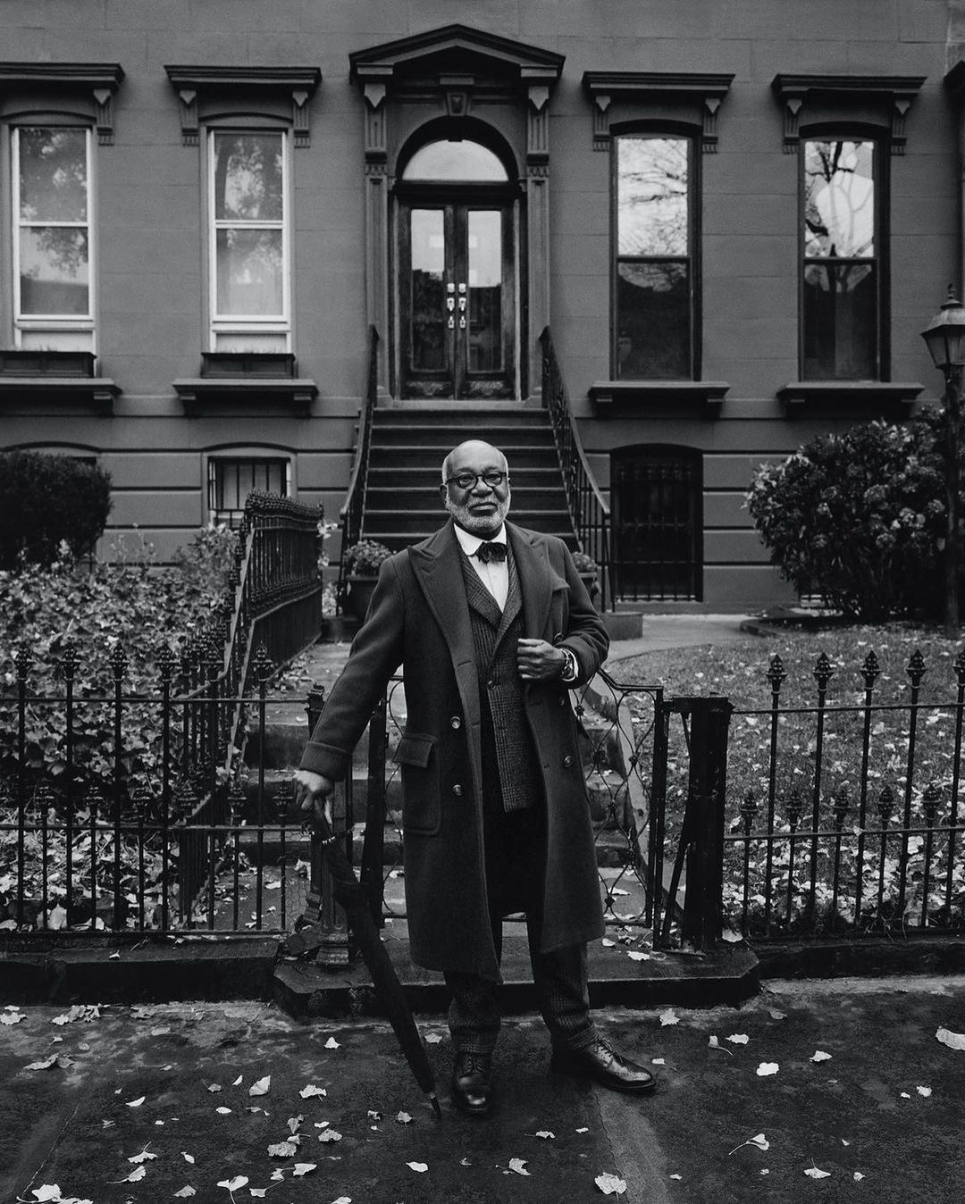 Monochrome portrait of an older adult male in a suit and coat, standing in front of a building with an umbrella, outdoors.
