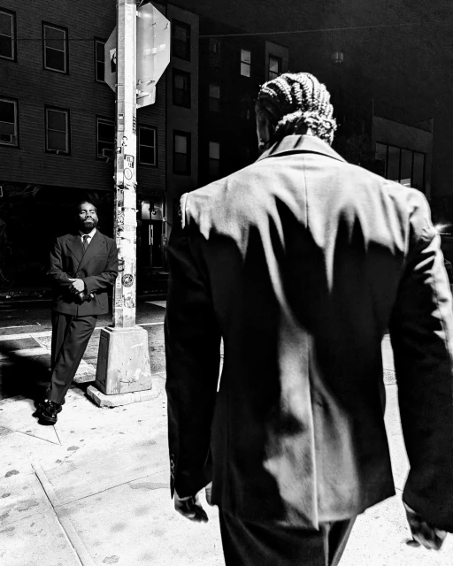 Two men in suits face each other on a city street at night in a black and white photograph. The man closer to the viewer has braids.