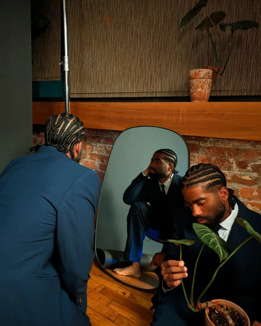 Man in a suit poses with a plant in front of a mirror reflecting his seated figure in an indoor setting, showcasing a stylish self-portrait.