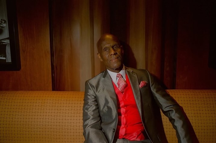 Elegant elderly African-American man in a suit and red vest sits on a couch in a dimly lit, wood-paneled room, exuding sophistication.