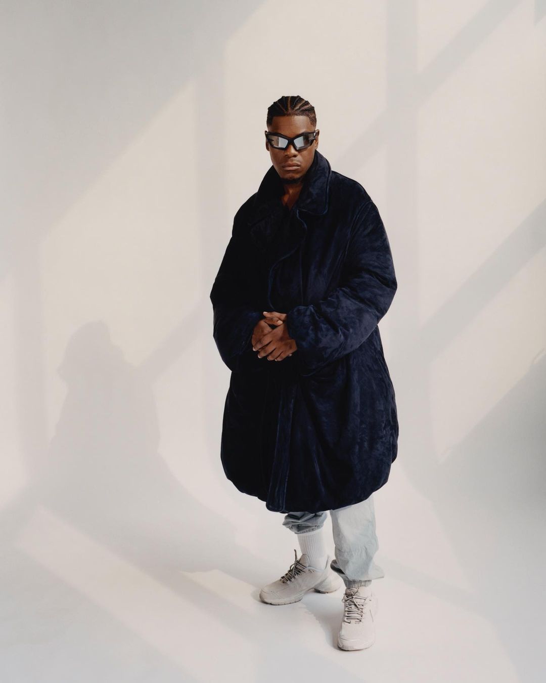 Person in dark coat, sunglasses, light jeans and sneakers in a studio with shadows and light.