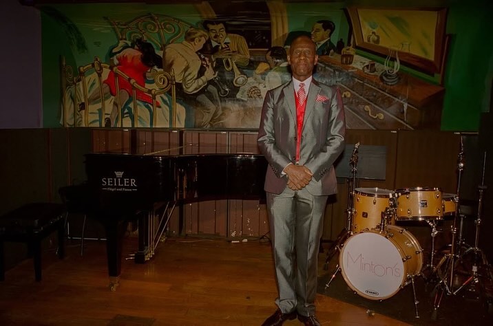 Adult male in a grey suit stands in front of a drum kit and piano in a dimly lit room with a jazz mural on the wall.