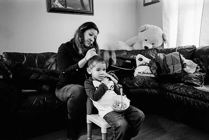 A mother braids her young child's hair in an indoor living room setting, the child sits on a small chair holding a bowl of food.