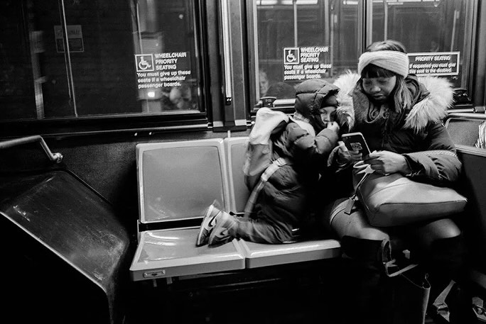 Black and white photo of an adult female and a child riding a bus; the woman looks at a smartphone while seated next to the child.