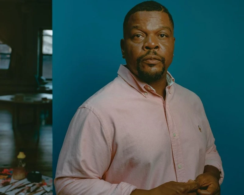 Close-up portrait of a Black man in a pink button-down shirt against a blue background, with a blurred room interior on the left.