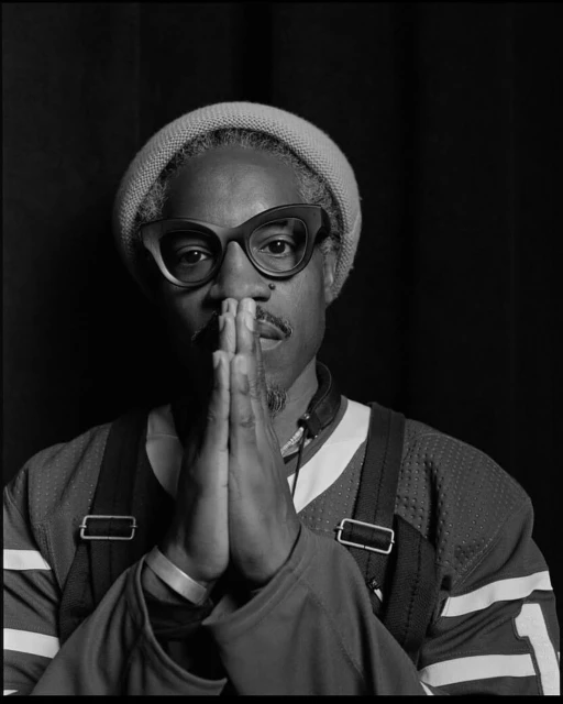 Black and white portrait of an adult Black man with glasses and a knit hat, hands clasped in front of a dark background.