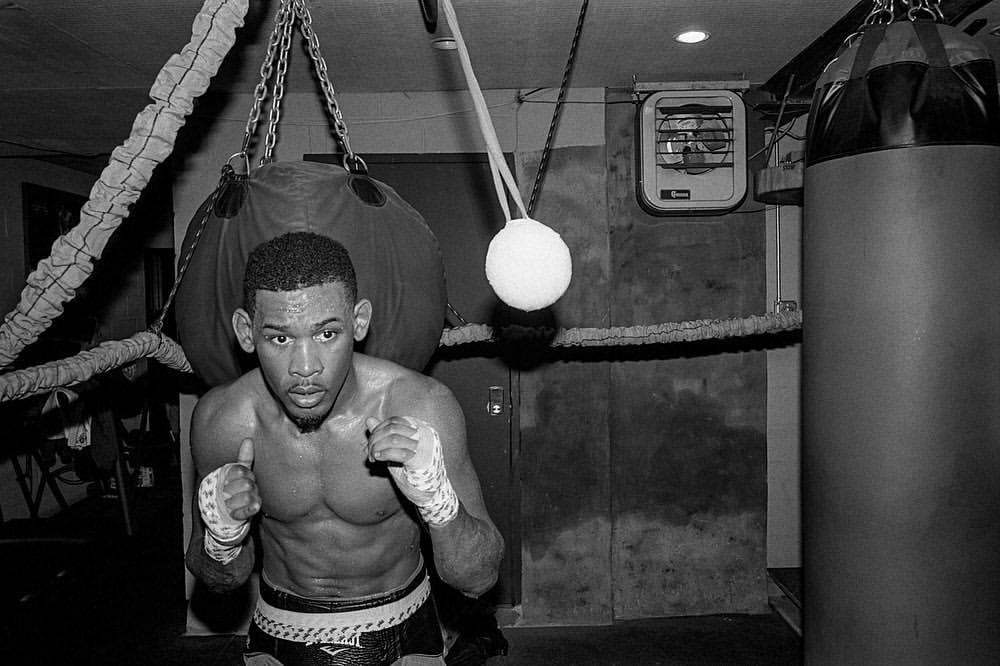 Black and white portrait of a muscular man with boxing wraps standing in a boxing gym with a heavy bag and speed bag.