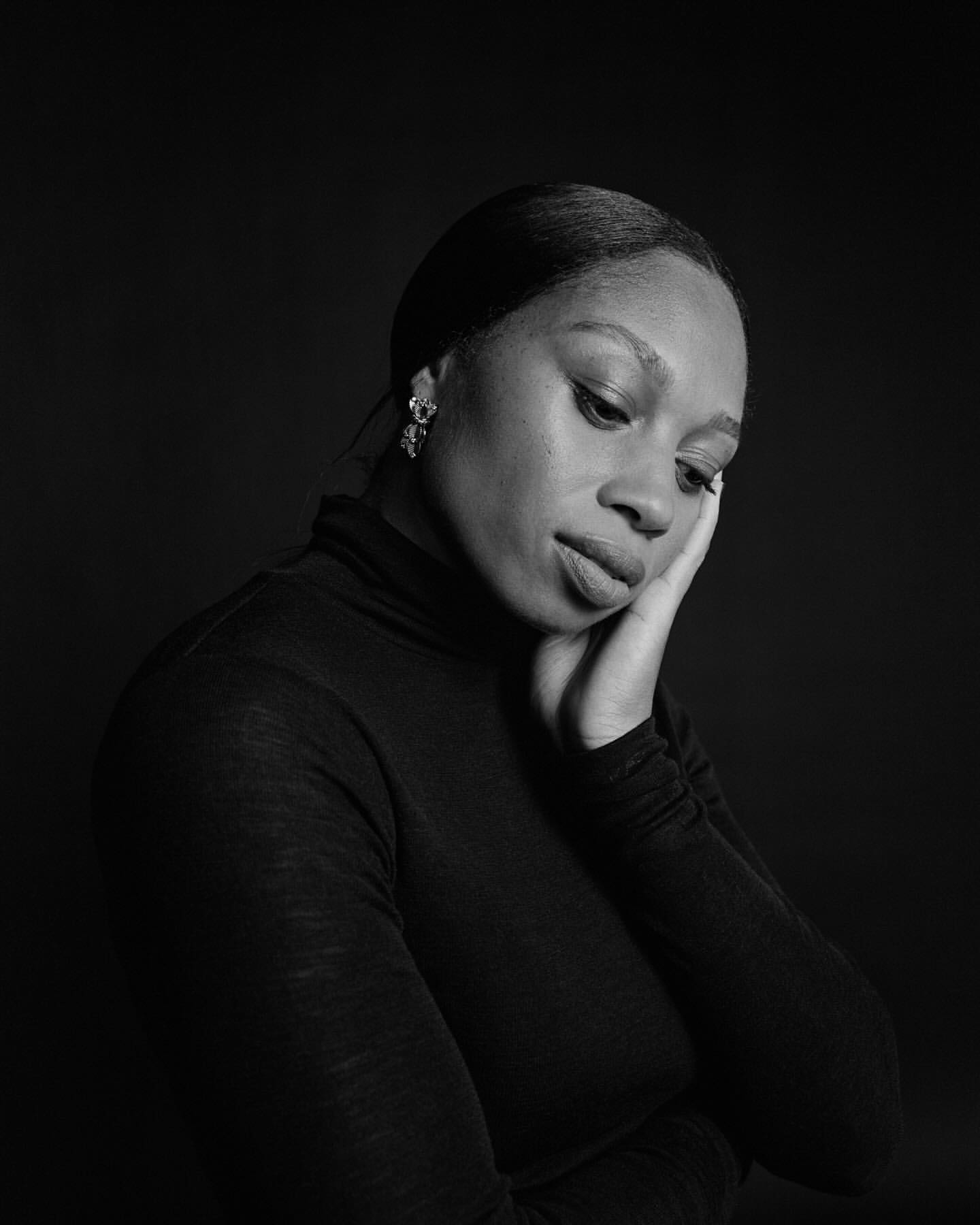 Black and white portrait of an adult woman with dark hair wearing a turtleneck sweater looking down and resting her face on her hand.