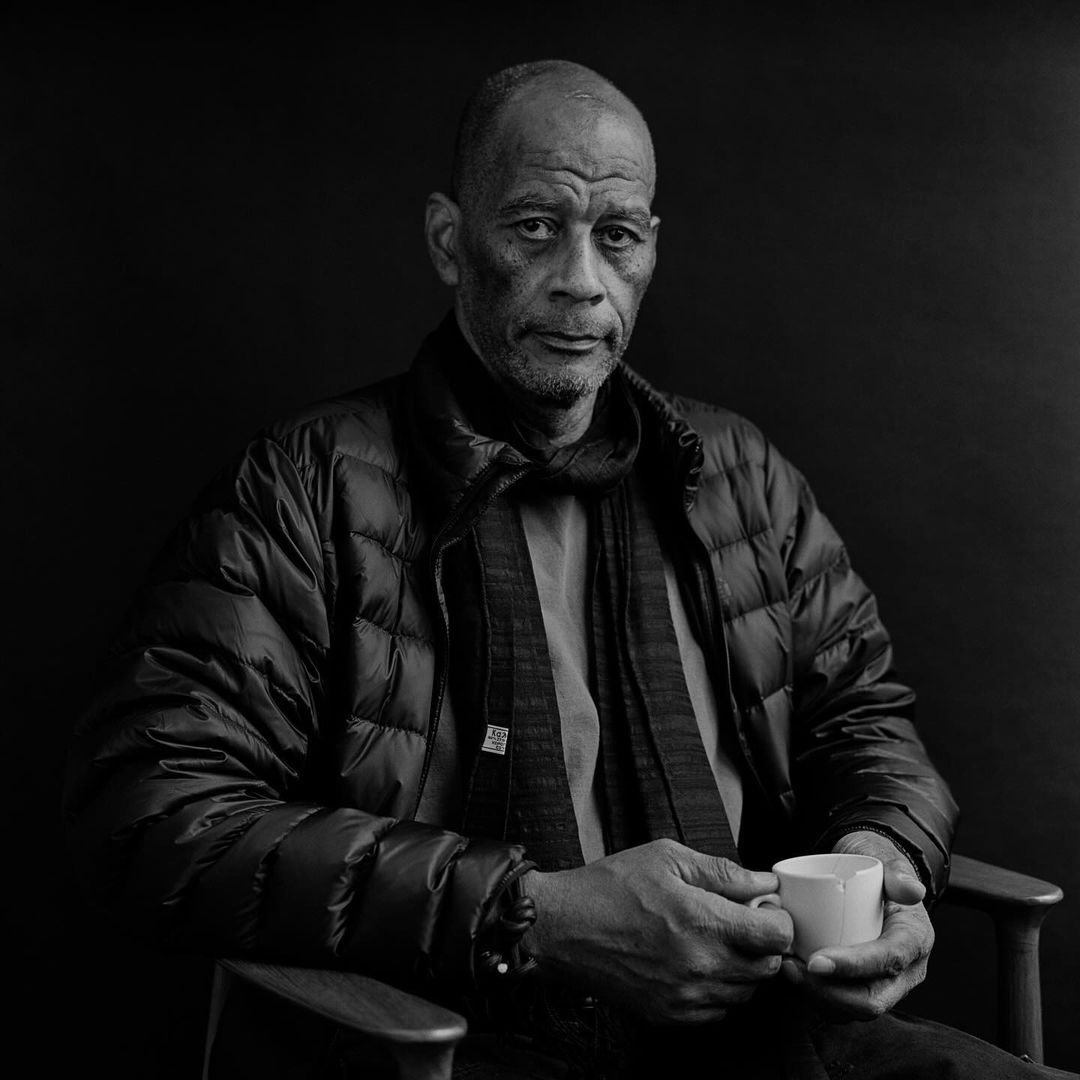 Black and white portrait of a mature African-American man in a puffer jacket, holding a cup, seated indoors against a dark backdrop.