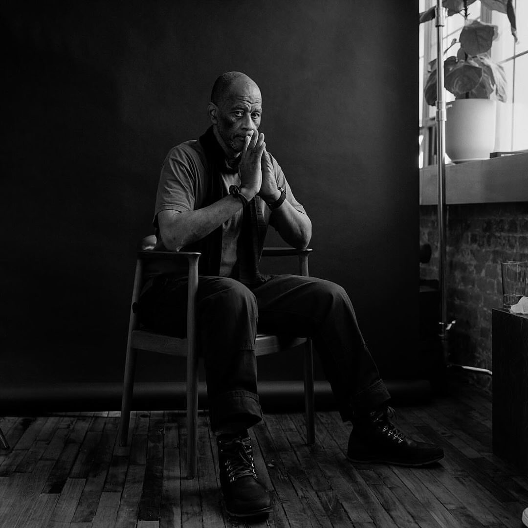 Black and white portrait of an African American man with his hands clasped sitting in a wooden chair in front of a dark background.