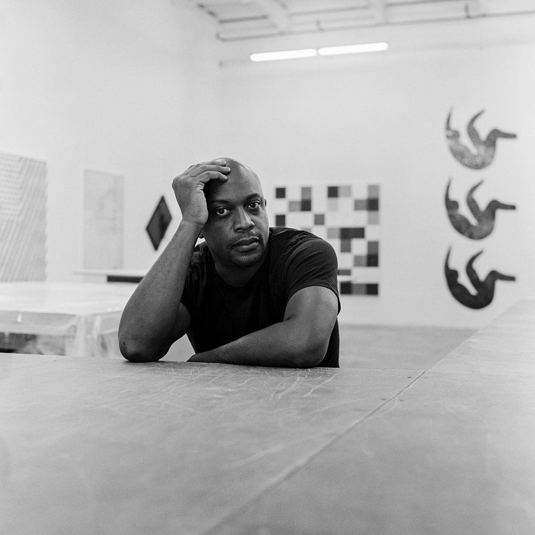 Portrait of a Black man in a black t-shirt leaning on a gray concrete surface, with various artworks on the white walls in the background.