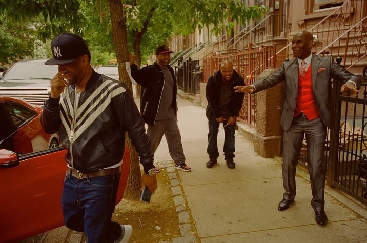 A group of four men stand on a sidewalk in front of an apartment building with trees, cars, and a red sports car on the side.