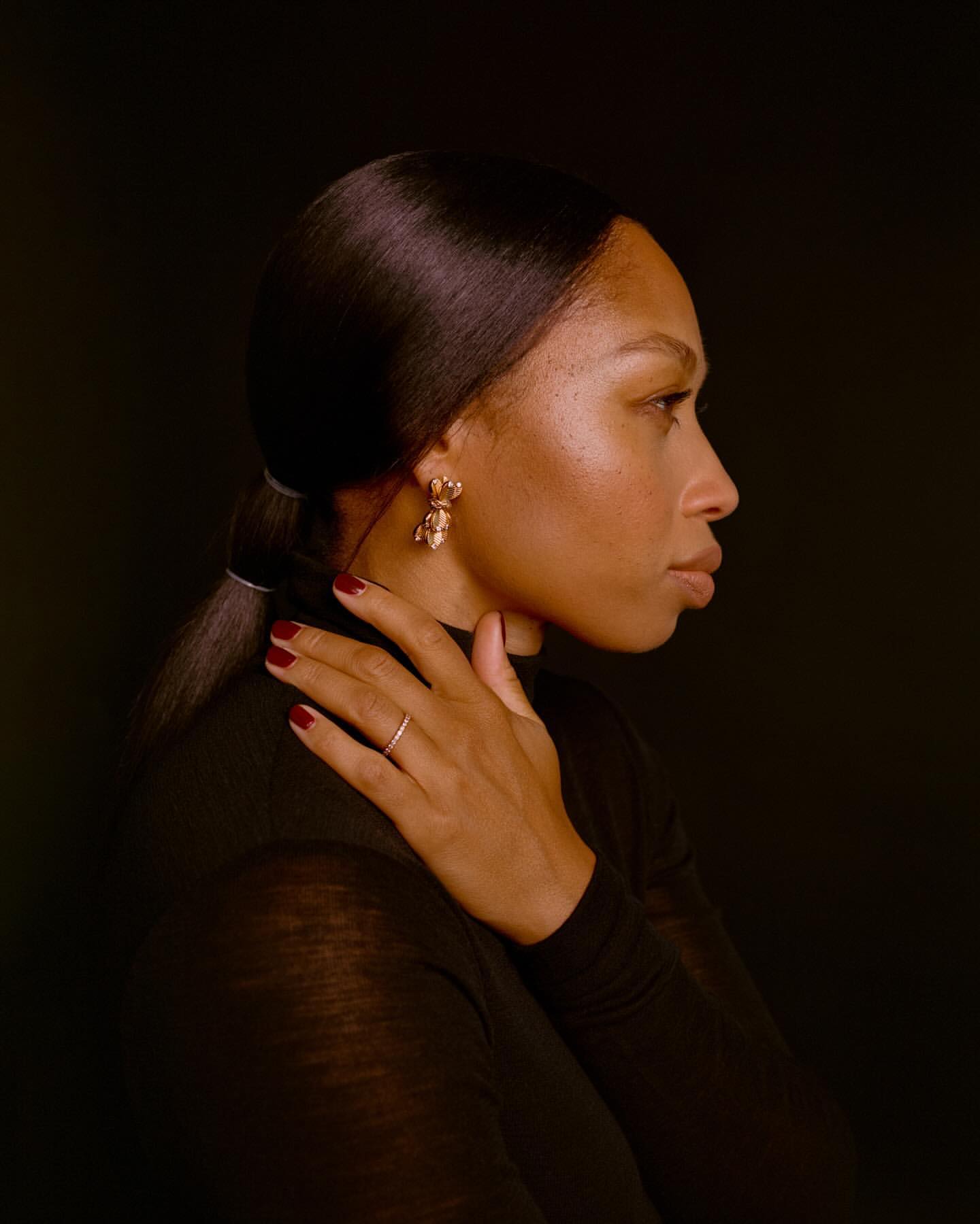 Profile portrait of a black woman in a black turtleneck touching her shoulder with dark red nails.