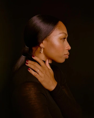 Profile portrait of a black woman in a black turtleneck touching her shoulder with dark red nails.