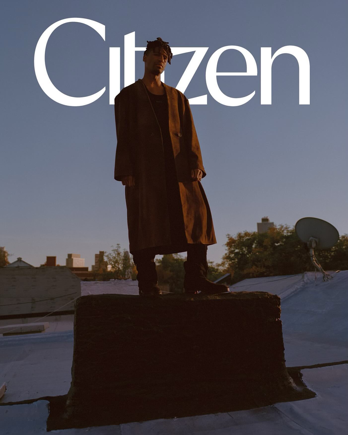 Man in coat stands on roof in urban scene, under 'Citizen' text. Outdoor shot with city buildings, clear sky at dusk or dawn.