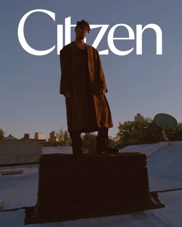 Man in coat stands on roof in urban scene, under 'Citizen' text. Outdoor shot with city buildings, clear sky at dusk or dawn.
