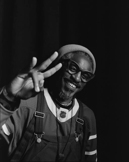 Black man in hockey jersey, glasses and beanie smiles and makes peace sign against dark background in portrait shot, looking at camera.