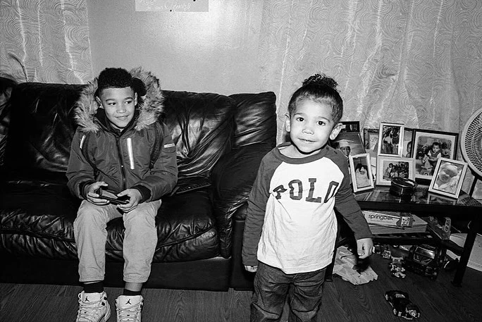 Two young boys indoors: one sits on a black leather couch playing a handheld game, while the other stands in front of a table.