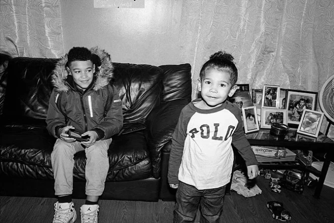 Two young boys indoors: one sits on a black leather couch playing a handheld game, while the other stands in front of a table.