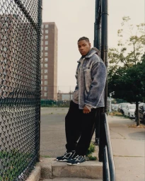 Man wearing a textured blue jacket and black trousers leaning against a chain-link fence in New York by Andre Wagner