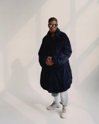 Man wearing dark sunglasses and an oversized dark coat standing against a light background with window shadows