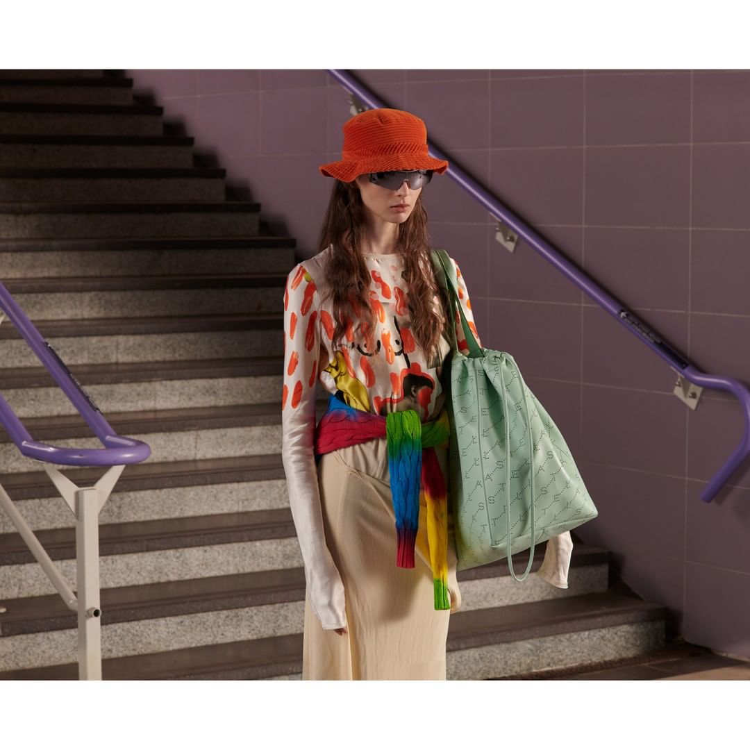 Woman wearing an orange bucket hat, patterned shirt, and holding a tote bag poses by a staircase in an indoor environment.