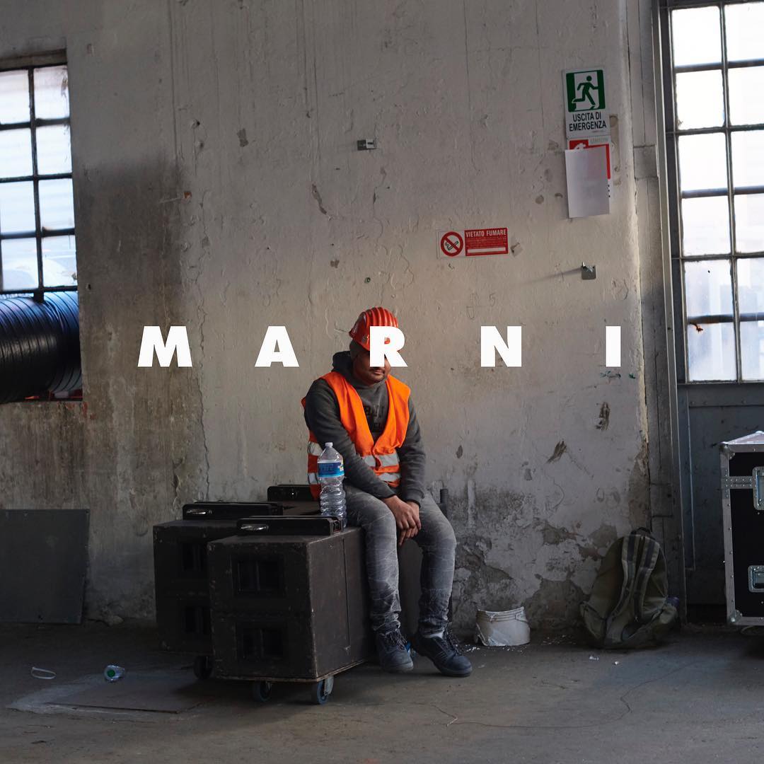 Man in hard hat and safety vest sits on speakers in an industrial space with a water bottle. Marni text overlay on the image.