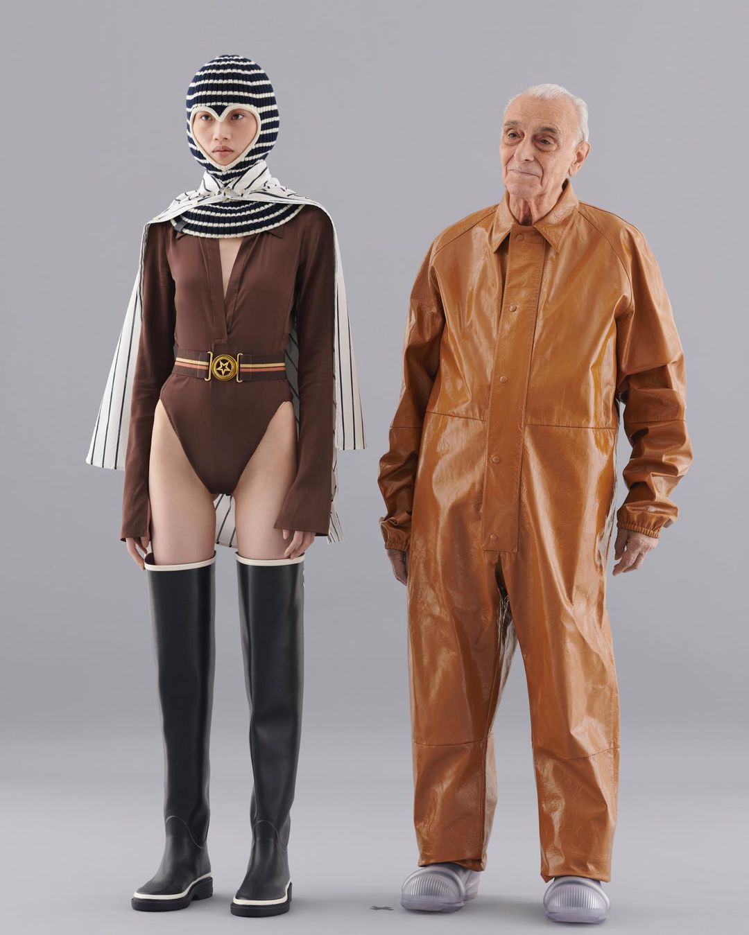 Fashion photo: Model in a brown bodysuit, striped head covering, and black boots stands next to an older man in a shiny brown jumpsuit.