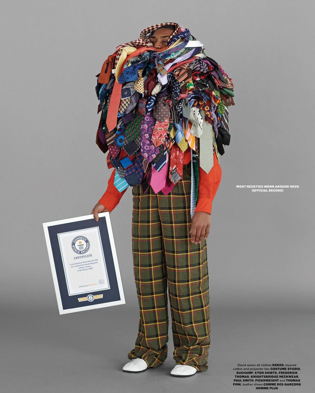 Man displays Guinness World Record certificate, wearing many neckties around his neck, wearing checkered pants, on a grey background.
