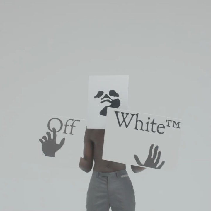 Torso shot of a man in grey pants holding Off-White branded cards with graphics in a sterile studio environment with even lighting.