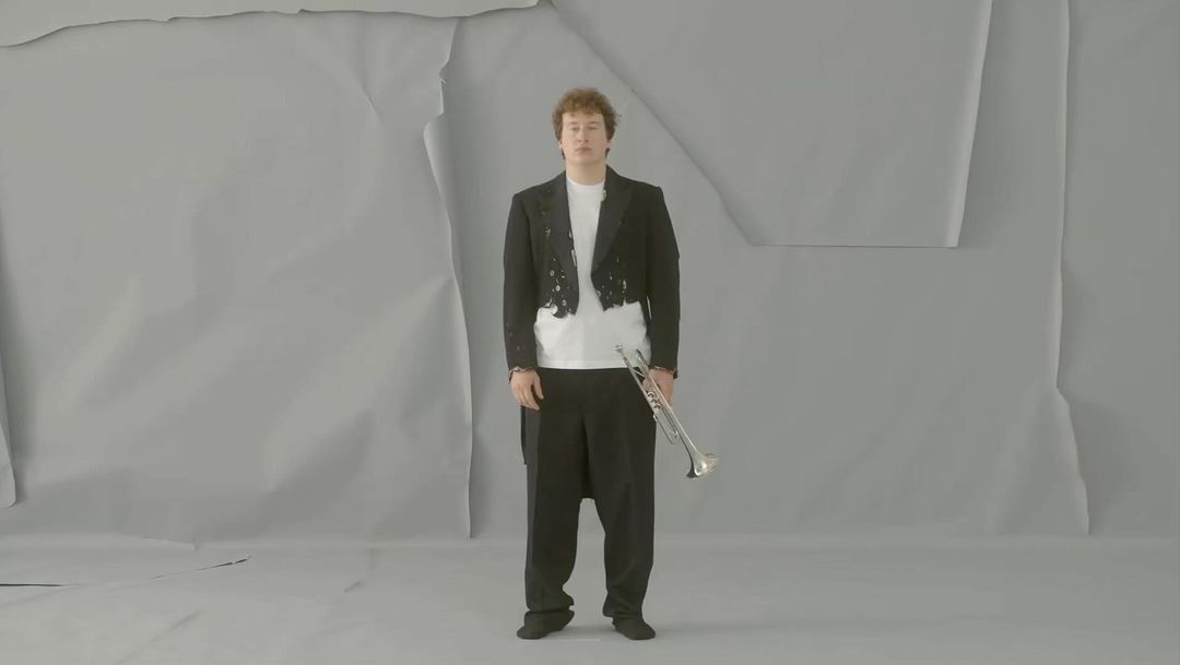 An adult male stands with a trumpet in front of a grey wrinkled paper background, wearing a black tuxedo jacket and black dress pants.