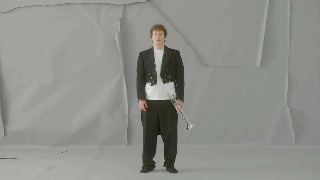 An adult male stands with a trumpet in front of a grey wrinkled paper background, wearing a black tuxedo jacket and black dress pants.