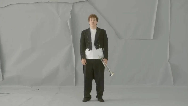 An adult male stands with a trumpet in front of a grey wrinkled paper background, wearing a black tuxedo jacket and black dress pants.