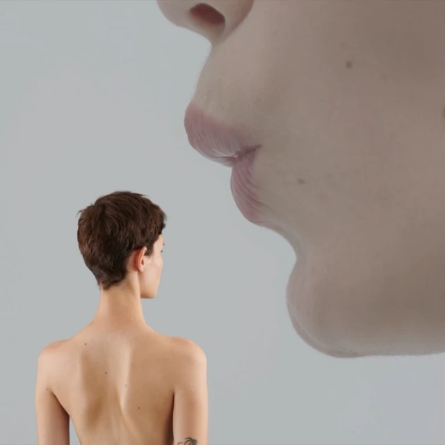 Surreal studio shot featuring an adult woman with short brown hair looking toward a floating, oversized face fragment against a gray background.