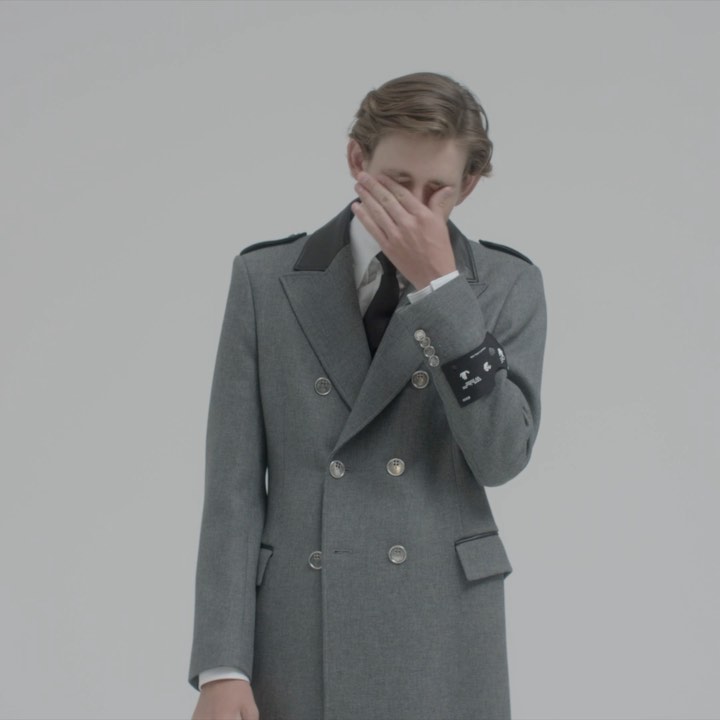Portrait of a stylish, light-skinned man in a grey coat with dark lapels and a tie, hand partially covering face, against white backdrop.