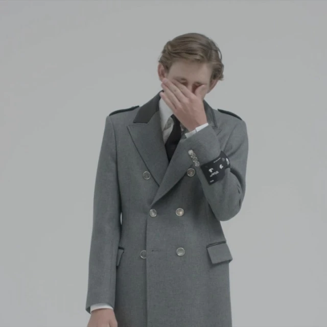 Portrait of a stylish, light-skinned man in a grey coat with dark lapels and a tie, hand partially covering face, against white backdrop.