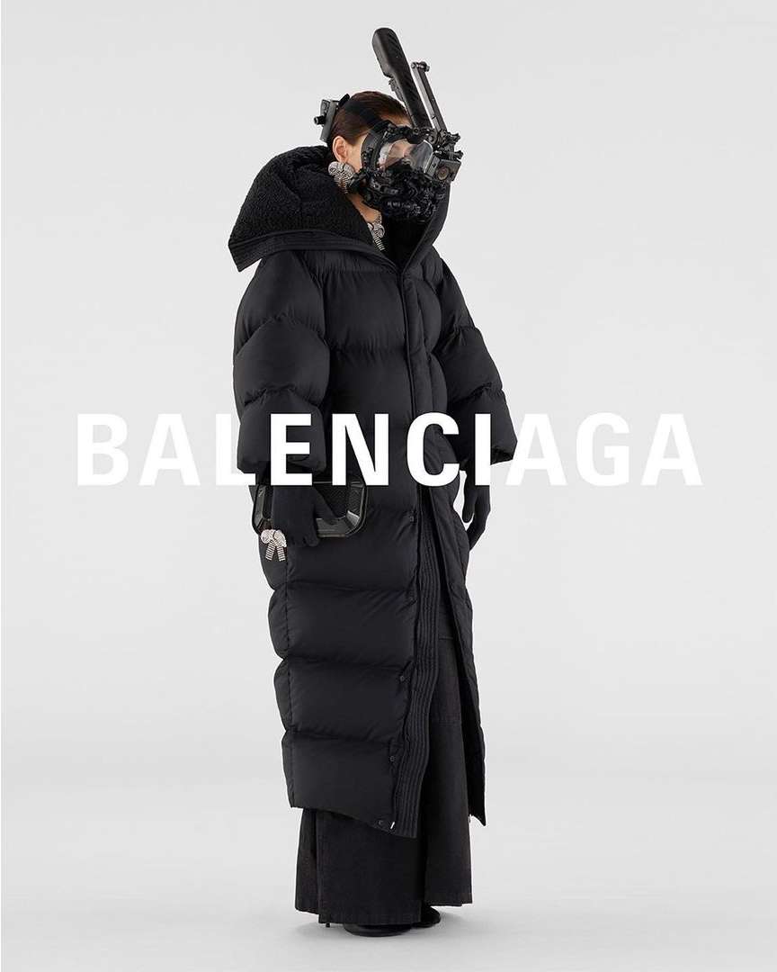 Fashion shot of a person wearing a large black puffer coat and a complex face mask with tubing in front of a bright white backdrop.