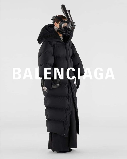 Fashion shot of a person wearing a large black puffer coat and a complex face mask with tubing in front of a bright white backdrop.