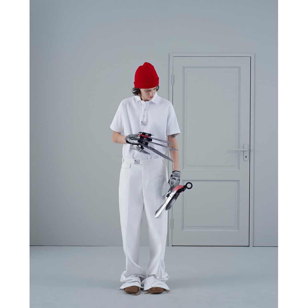 Person in white outfit and red beanie holding two unique bladed objects in a light grey photo studio with a door in the background.