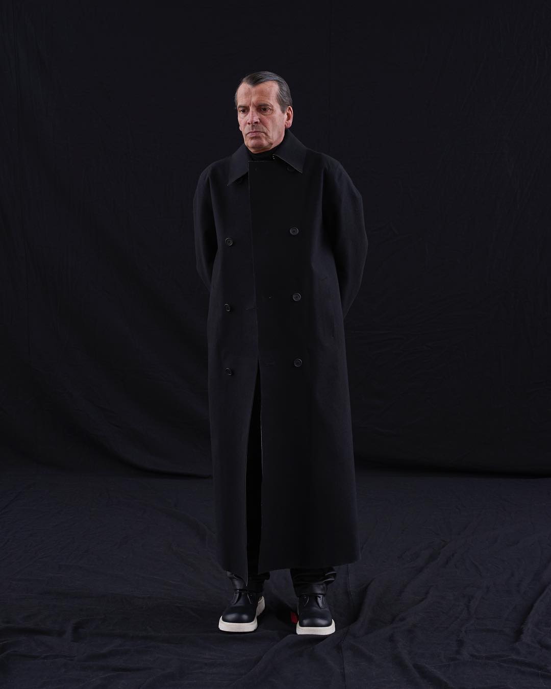 Full shot of a man in a black, double-breasted overcoat posing against a dark background in black pants and white-sole shoes.