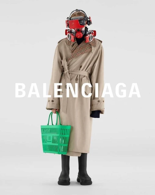 A person wearing a beige trench coat, black boots, and a red futuristic helmet holds a green shopping basket in a fashion setting.