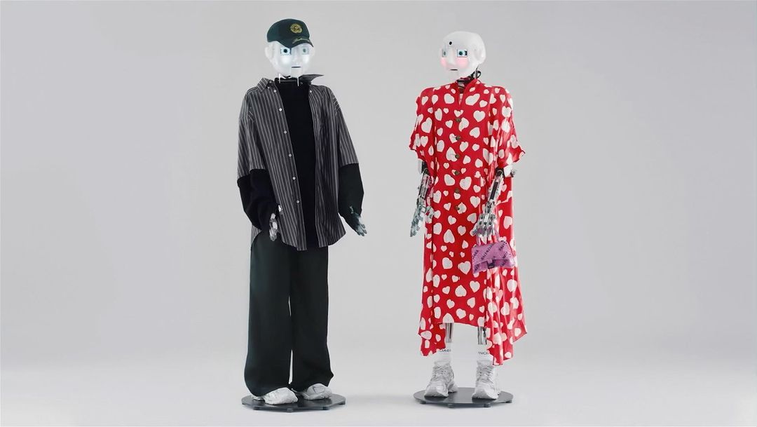 Two humanoid robots are styled in contemporary fashion, standing in a bright studio. One is in streetwear and the other in a patterned dress.