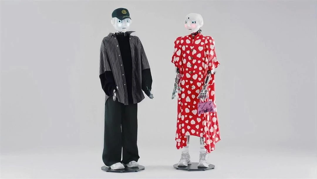 Two humanoid robots are styled in contemporary fashion, standing in a bright studio. One is in streetwear and the other in a patterned dress.