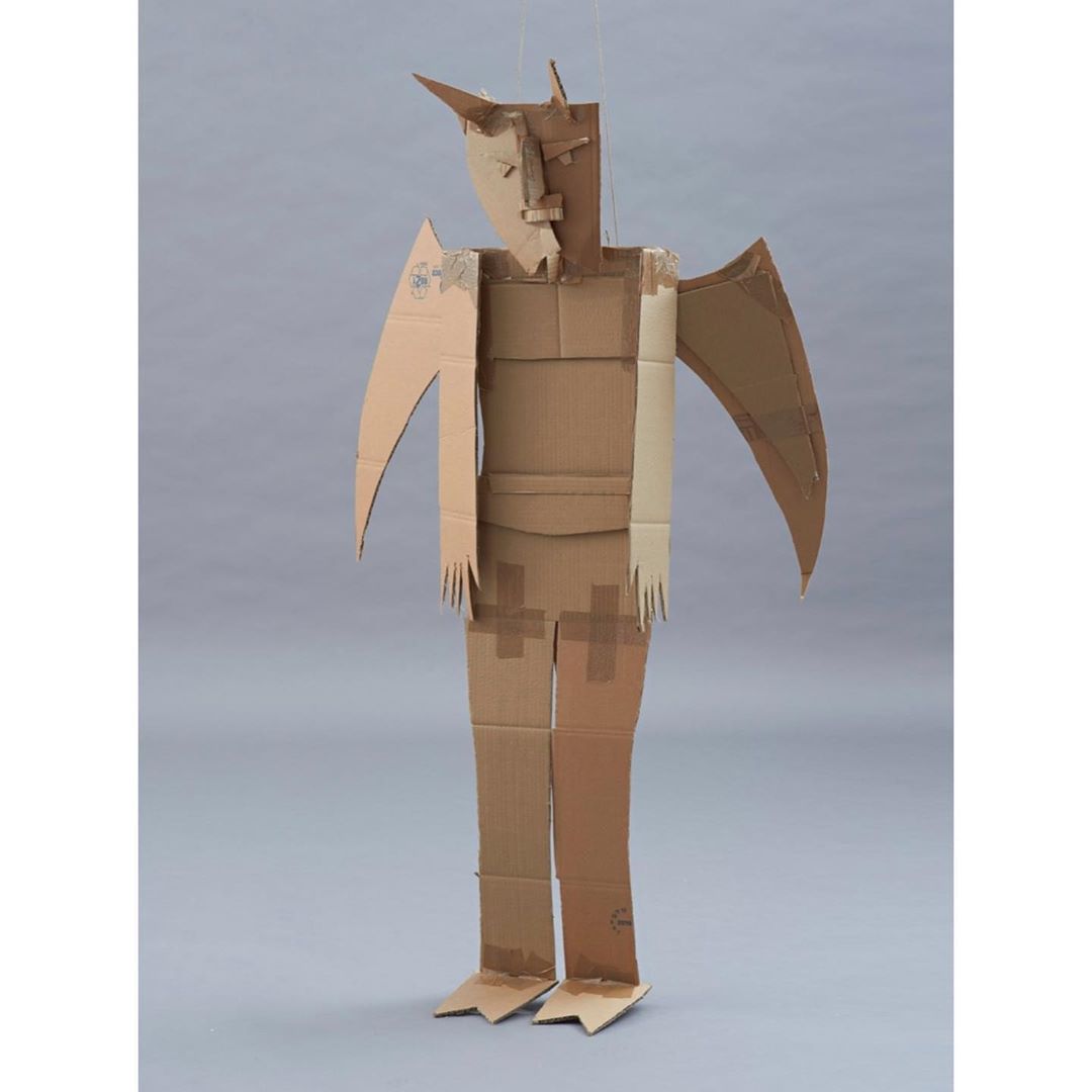 Full shot of a humanoid cardboard sculpture with wing-like arms standing in a photo studio against a gray backdrop.