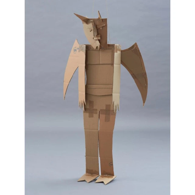 Full shot of a humanoid cardboard sculpture with wing-like arms standing in a photo studio against a gray backdrop.