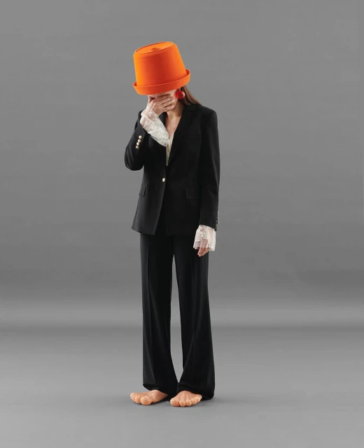Adult female fashion model in a black business suit with lace cuffs wearing a bright orange hat covering her eyes and bare feet on gray.