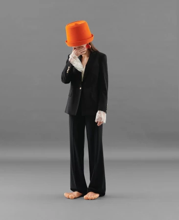 Adult female fashion model in a black business suit with lace cuffs wearing a bright orange hat covering her eyes and bare feet on gray.