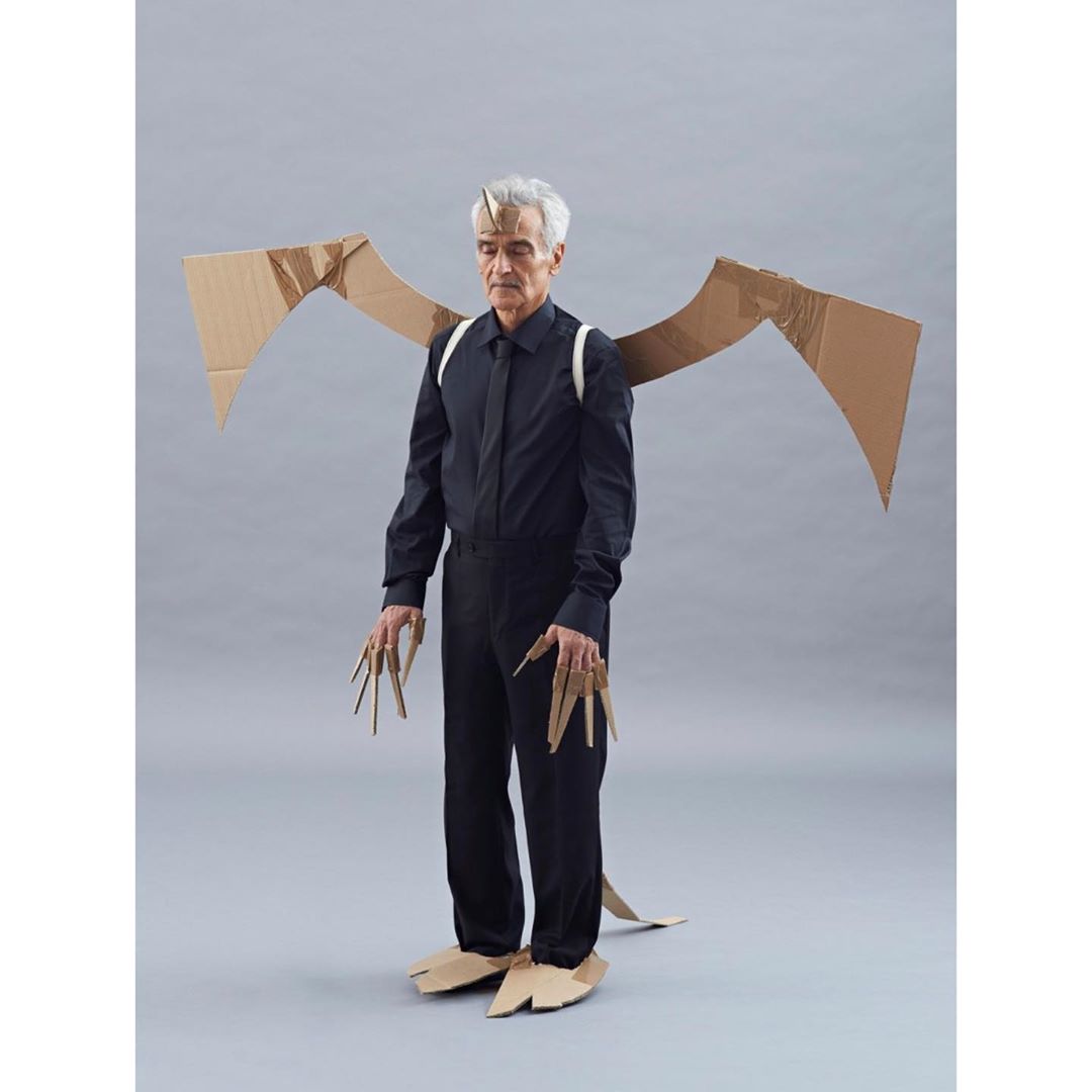 Full shot of a man in a black shirt and pants wearing a cardboard dragon costume with wings, horns, claws, and feet against a grey backdrop.